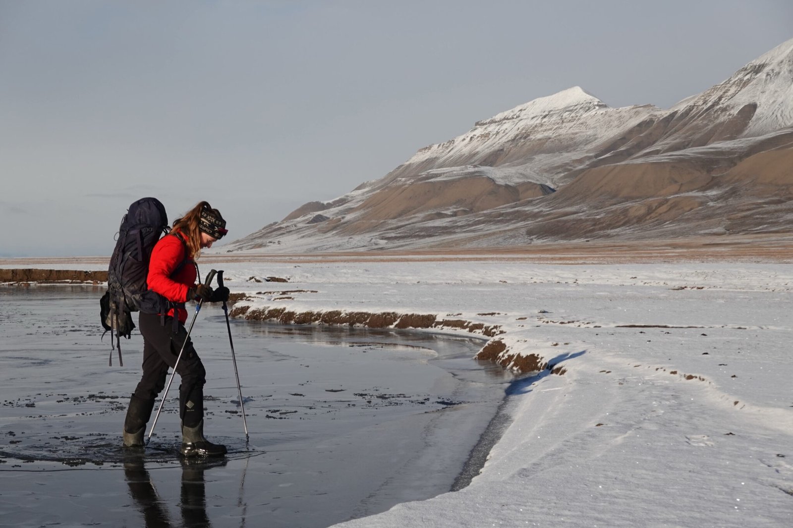 A student with trekking poles wades across a shallow river in early snow